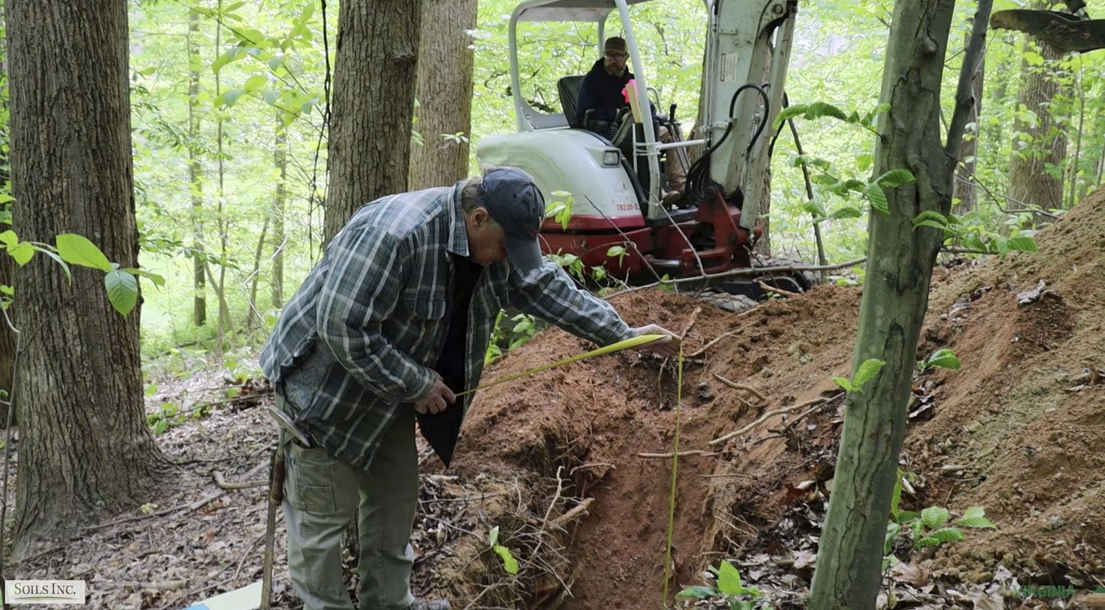 A Worker From Soils Inc Standing At A Dug Hole Holding A Measure To Inspect The Depth With A Tractor Parked Behind Him