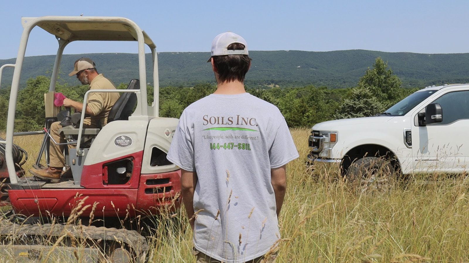 A Back Shot Of A Man In The Branded Shirt Of Soils Inc Looking After The Tractor Working On The Soil Inspection