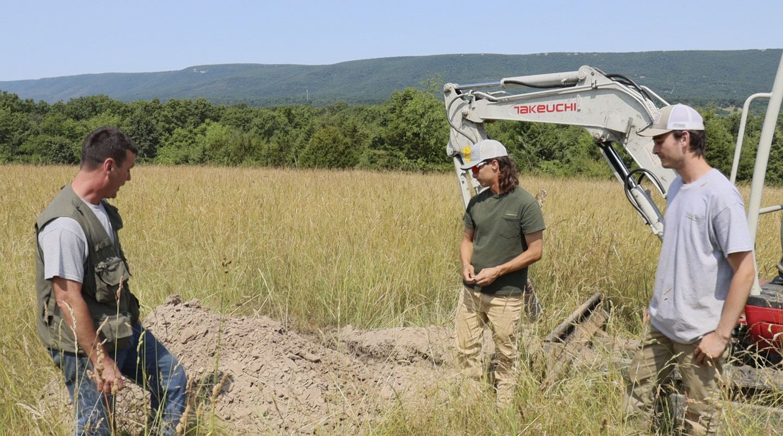 Three Soil Professionals From Soils Inc Are Inspecting The Dug Out Soil In A Grassy Area With A Tractor Parked Behind Them